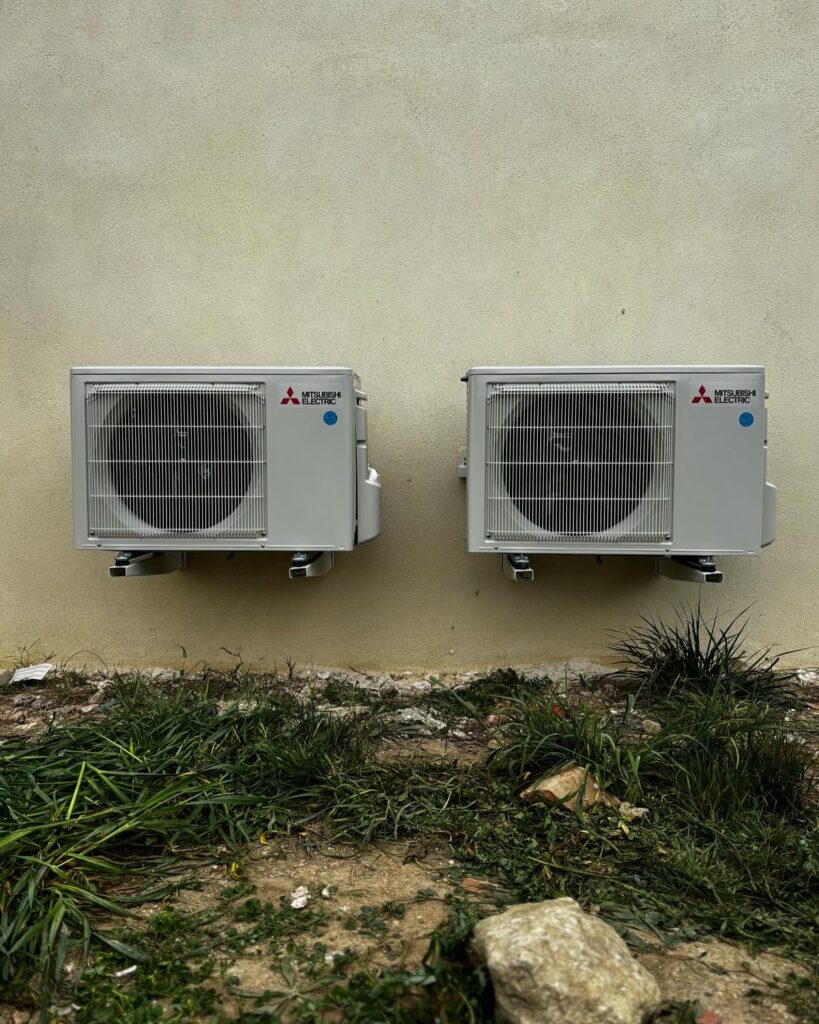 Deux unités extérieures de climatisation Mitsubishi Electric fixées sur un mur beige au-dessus d'une zone envahie par l'herbe.