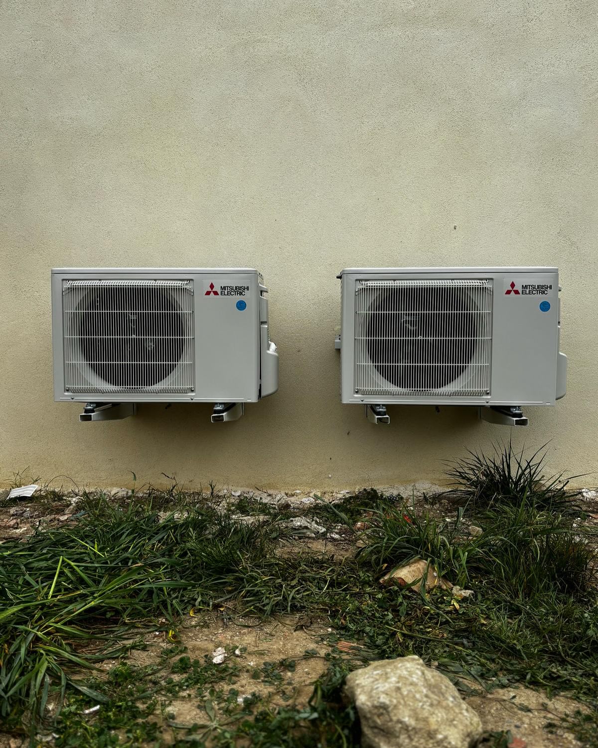 Deux unités extérieures de climatisation Mitsubishi Electric fixées sur un mur beige au-dessus d'une zone envahie par l'herbe.
