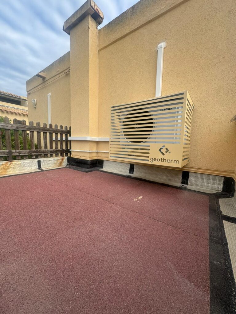Unité extérieure de pompe à chaleur Geotherm Bosch sur une terrasse avec sol rouge, fixée contre un mur jaune et près d'une souche de cheminée.