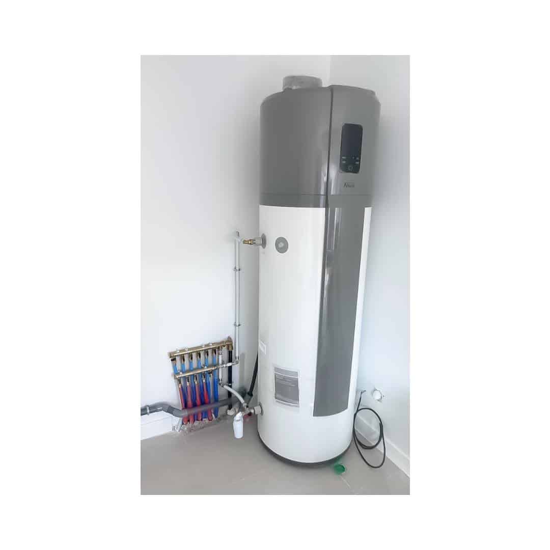 Ballon d'eau chaude thermodynamique blanc et gris avec panneau de contrôle affichant 'Aira', près d'un collecteur de chauffage au sol avec tuyaux rouges et bleus.