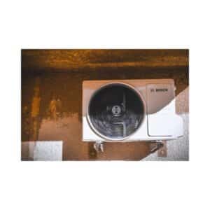 Unité extérieure de climatiseur blanc Bosch fixée à un mur texturé et ocre, avec un ventilateur visible derrière la grille.