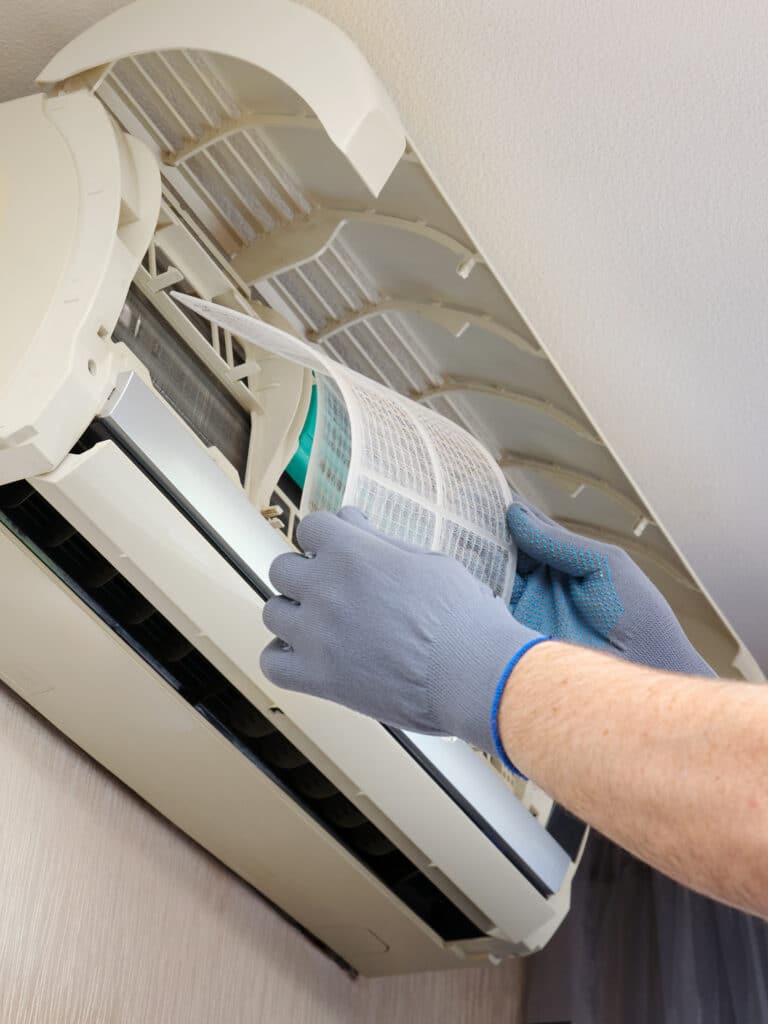 Mains gantées retirant un filtre à air sale d'une unité intérieure de climatiseur mural pour nettoyage.
