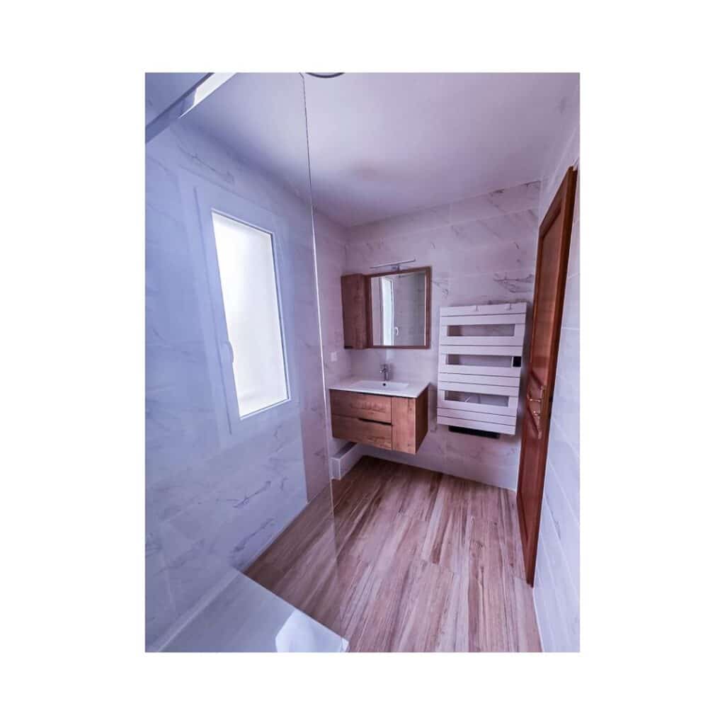 Salle de bain moderne avec douche vitrée, meuble vasque en bois flottant, miroir, et sèche-serviettes mural blanc.