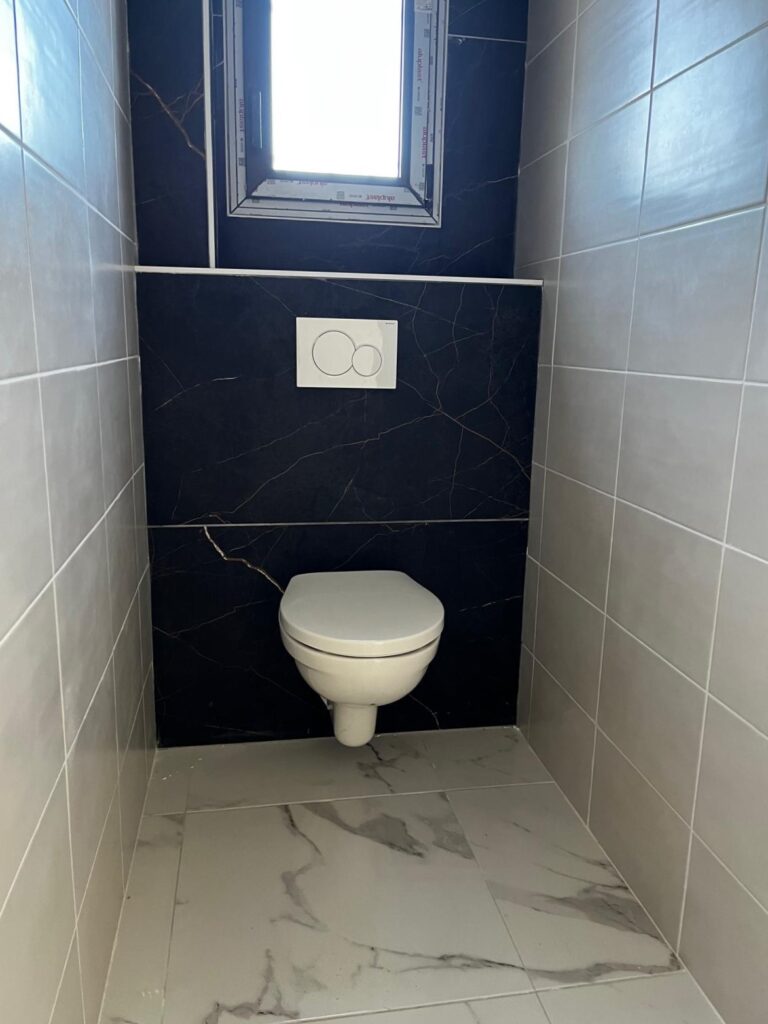 Toilette suspendue dans un WC moderne, murs carrelés gris et panneau mural noir marbré avec boutons de chasse d'eau Geberit.