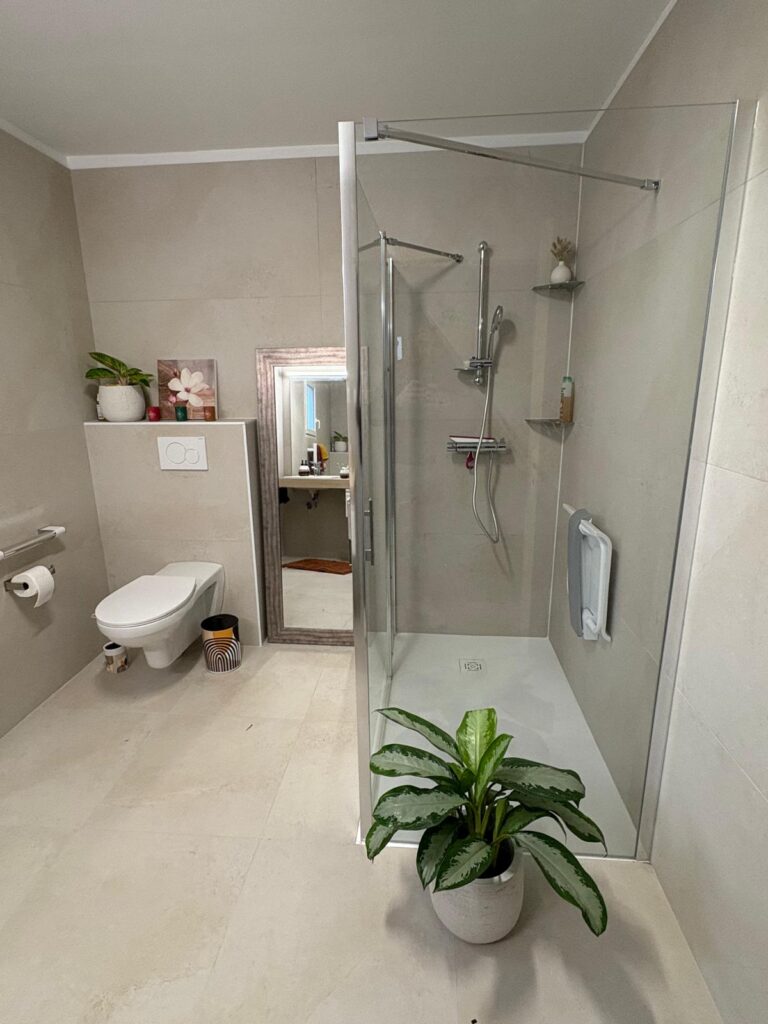 Salle de bain beige et moderne avec douche à l'italienne, WC suspendu et grand miroir. Décoration naturelle, plante verte, et barre d'appui.